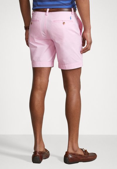 Roza bombažna kratka hlače s prilagojenim krojem, ki vključujejo zadnje žepke in gumbne detajle. V kompletu z rjavimi usnjenimi čevlji.