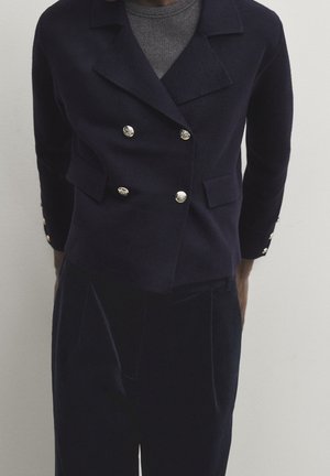 Navy double-breasted jacket in textured wool with metallic buttons, featuring a notched collar and two front pockets. Paired with dark velvet trousers.