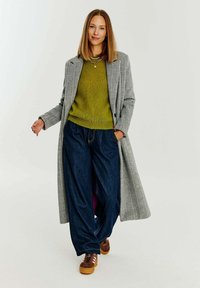 Dlouhý kabát s houndstooth vzorem, olivově zelený svetr, široké džíny a hnědé tenisky se žlutou podrážkou, které nosí žena stojící vzpřímeně.