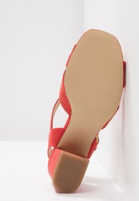 Sandale en daim rouge à talon bloc, semelle texturée couleur caramel, et bout ouvert, positionnée semelle vers le haut contre un mur d'angle blanc.