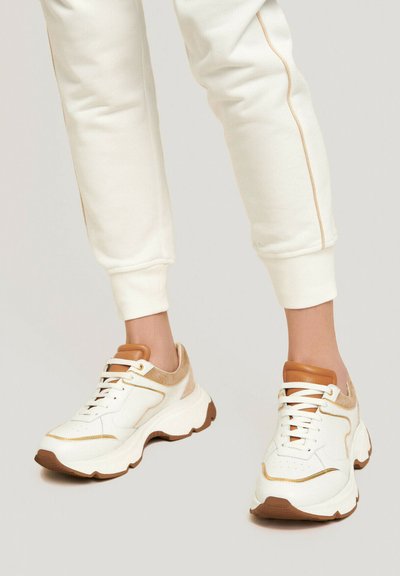 Zapatillas blancas con acentos beige, suela gruesa, material texturizado y tracción marrón en contraste. Los cordones son blancos y cuentan con una lengüeta reforzada.