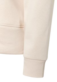 Hellbeige Sweatshirtstoff im Close-up, mit einem gerippten Bündchen und glatter Textur, in einem einfachen, minimalistischen Design.