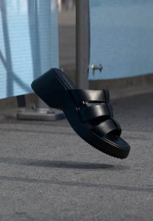 Must platvormsandaal kolme laia rihmaga hõljub halli kõnnitee kohal siniste kangasplaatide lähedal.