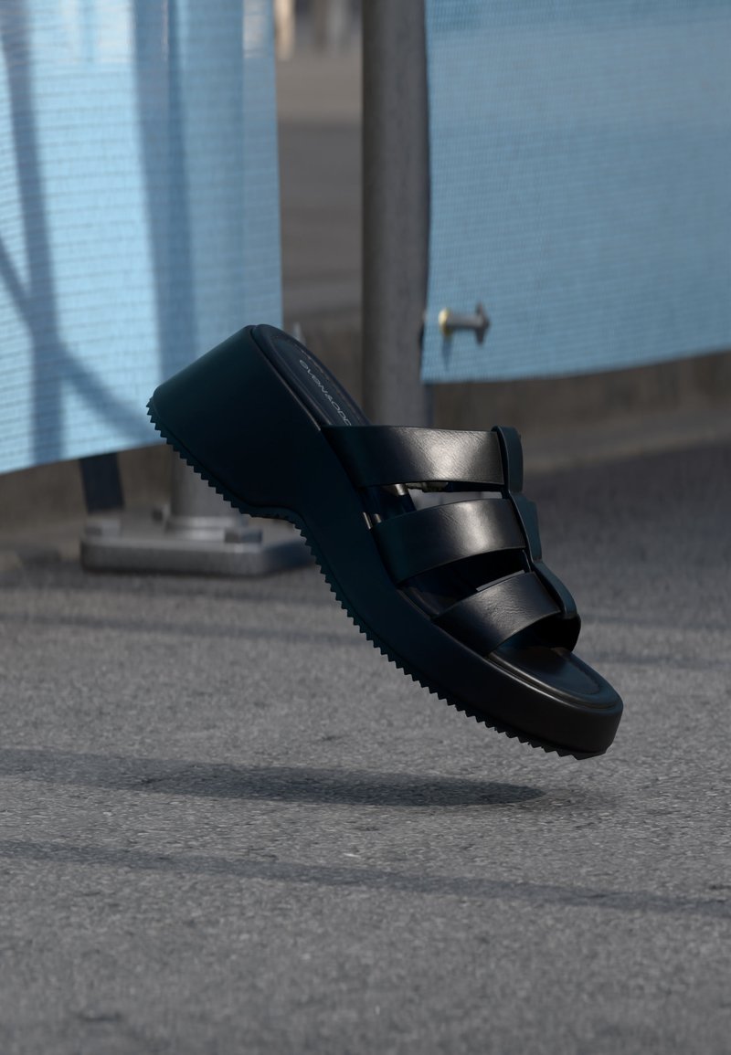 Μαύρο σανδάλι πλατφόρμα με τρεις φαρδιές λουρίδες που αιωρείται πάνω από γκρι πλακόστρωτο κοντά σε μπλε υφασμάτινες επιφάνειες.