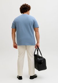 Άνδρας φαινομενικά από πίσω, φορώντας μπλε πουκάμισο με κοντά μανίκια, μπεζ παντελόνι, μαύρα παπούτσια, κρατώντας μια μαύρη δερμάτινη τσάντα ταξιδίου στο δεξί του χέρι.