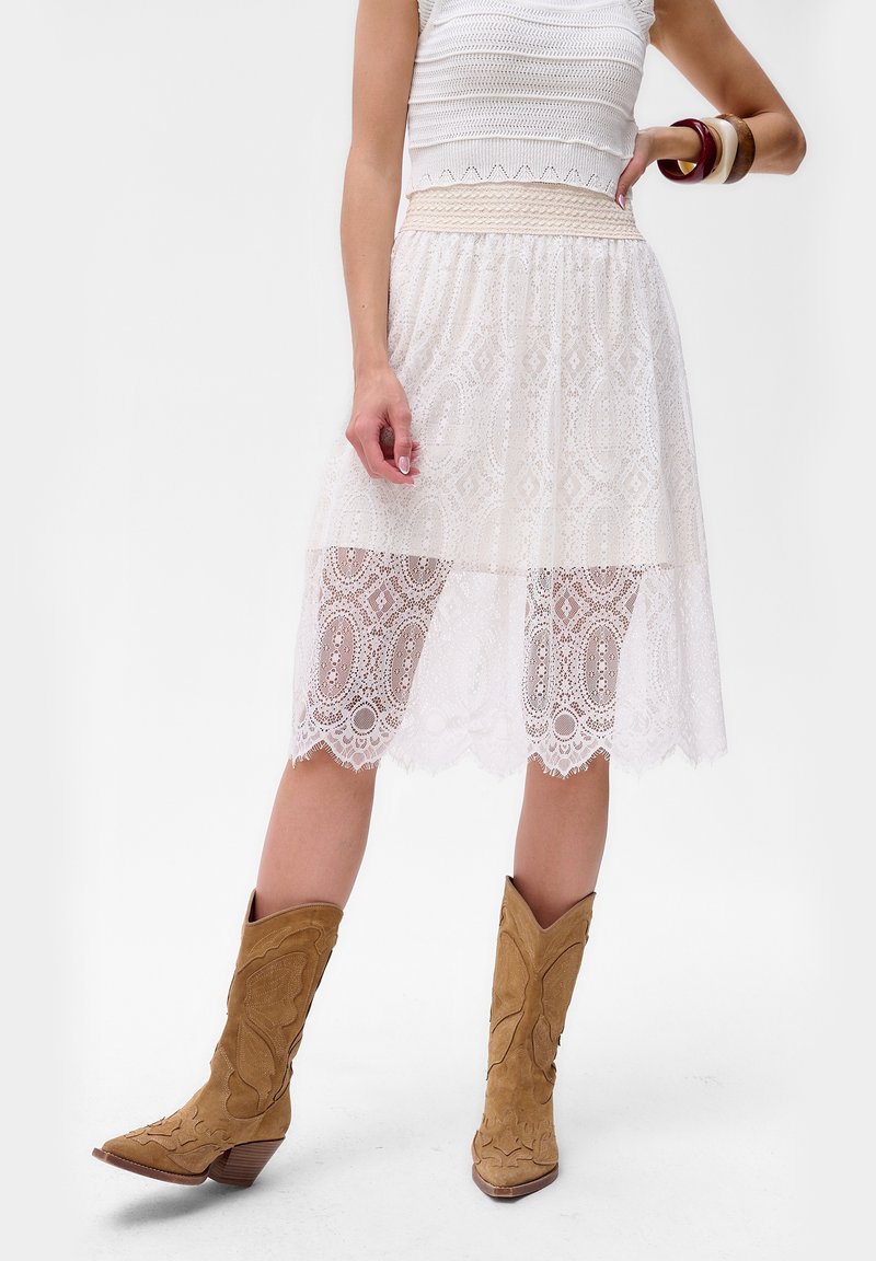 Jupe en dentelle blanche avec une couche inférieure transparente, présentant des motifs complexes et une taille texturée. Associée à des bottes de cowboy beige.