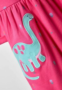 Ružová látka s malými kvetinovými potlačami a veľkým svetlo modrým dinosaurím applique so š pinky bodkami zošitými na nej.