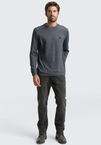 Graues Strick-Oberteil mit langen Ärmeln und rundem Halsausschnitt, kombiniert mit dunkelblauen Jeans und grauen Stiefeln, mit einem kleinen Logo-Detail auf der Brust.