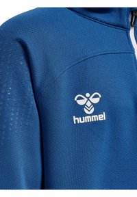 Blå idrottsjacka i lättviktigt material, med ett strukturerat mönster på axeln och en vit logotyp på bröstet.