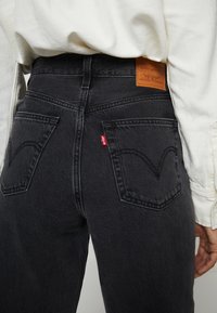 Jeans de mezclilla negra de cintura alta, con bolsillos traseros que presentan un patrón de costura clásico y una etiqueta de cuero marrón en la cinturilla.