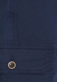Primo piano di un tessuto blu navy che mostra una tasca cucita con un bottone di legno. Texture liscia e linee pulite con le cuciture dettagliate visibili.
