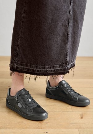 Black sneakers feature a mix of smooth leather and textured mesh, with laces and a side zipper accent. The wearer has frayed black denim skirt.