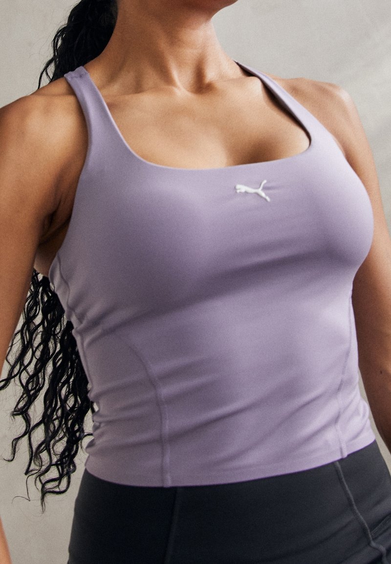 Femme portant un débardeur de sport ajusté lavande avec un logo Puma et des leggings de sport noirs, cheveux longs et bouclés visibles de dos.