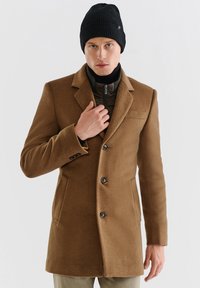 Brązowy wełniany płaszcz z trzema guzikami, klapami w kształcie notcha oraz bocznymi kieszeniami. Noszony na czarnej kurtce zapinanej na zamek i czarnej czapce z prążkowanej wełny.