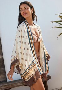 Kimono léger en crème avec des motifs floraux et géométriques, bord bleu et accents marron ; design à ouverture frontale et manches amples.