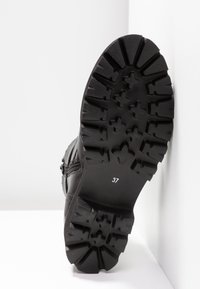 Semelle de botte noire avec des crampons profonds et le numéro de taille visible 37, positionnée verticalement contre un mur et un sol blancs.