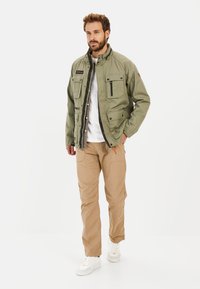 Olive green jacket with multiple pockets, zip closure, and high collar, paired with beige pants and white sneakers.