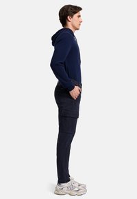 Felpa con cappuccio navy, pantaloni cargo slim-fit e sneakers bianche. I pantaloni presentano tasche laterali e una texture in tessuto liscia e resistente.