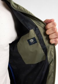 Interno della giacca verde oliva con rivestimento nero caratterizzato da un motivo a logo ripetuto; include una tasca sinistra con un'etichetta blu e bianca.