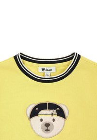 Camiseta amarilla con un cuello a rayas en blanco y negro, que presenta un oso bordado con una gorra en el pecho. Etiqueta visible dentro del cuello.