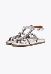 Sandali metallici argento con gemme rotonde decorative sulle cinghie, suola piatta e fibbia per caviglia regolabile. Texture liscia con design minimal.
