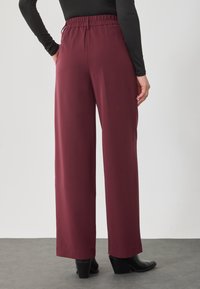 Kobieta ubrana w bordowe, szerokie spodnie z wysokim stanem i elastycznym paskiem, w połączeniu z czarnymi botkami sięgającymi kostek oraz czarną bluzką z długim rękawem.