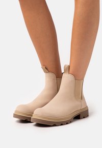 Beige kotníkové boty vyrobené z hladkého materiálu, s elastickými bočními panely, poutky na zadní straně a texturovanou gumovou podrážkou s hlubokým vzorkem.
