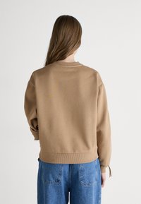 Brown sweatshirt with a crew neckline, dropped shoulders, and ribbed cuffs, worn with blue loose-fitting jeans. Smooth fabric texture.