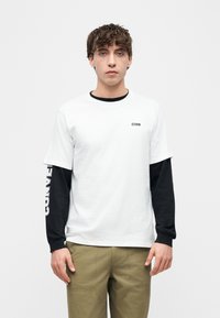 Νεαρός άνδρας με λευκό κοντομάνικο μπλουζάκι CONVERSE φορεμένο πάνω από μαύρο μακρυμάνικο και χακί παντελόνι, στέκεται μπροστά σε ένα απλό φωτεινό φόντο.