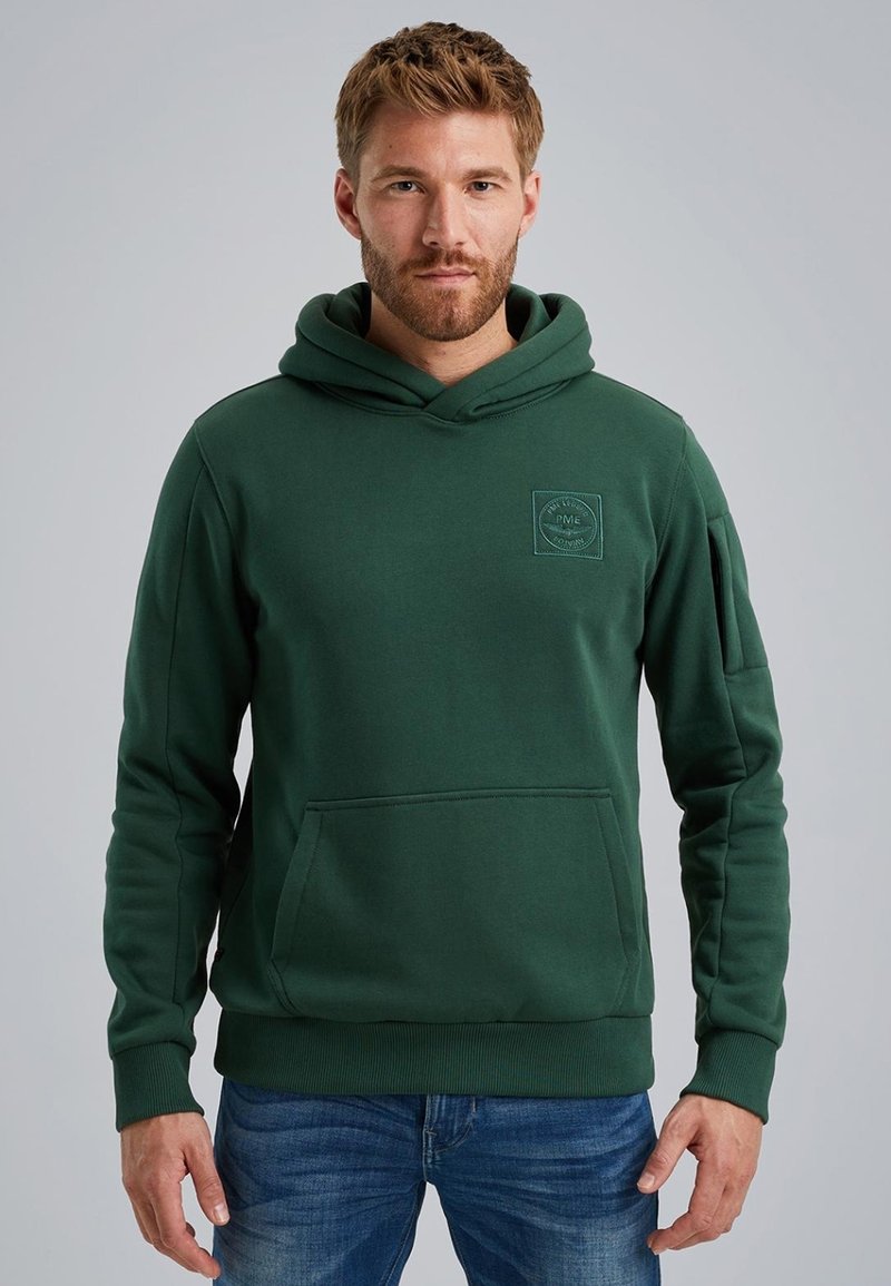 Green hoodie made of cotton-blend material, featuring a kangaroo pocket, ribbed cuffs, and a patch logo on the chest. Casual design.