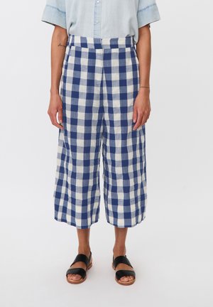 Pantalon large court à carreaux bleus et blancs porté avec une chemise bleu clair à manches courtes boutonnée et des sandales noires.