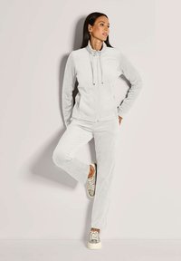 Vrouw in een lichtgrijze zip-up jas en bijpassende broek staat tegen een muur, met een gebogen been, en draagt patroon sneakers.