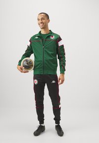 Chaqueta atlética verde con cremallera, con acentos en negro y rojo, combinada con pantalones de chándal negros que llevan un logo y franjas laterales rojas. Sosteniendo un balón de fútbol.