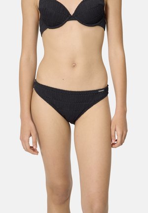 Mujer con un conjunto de bikini negro texturizado, con un top estilo sujetador y braguitas de tiro bajo, sobre un fondo claro y liso.