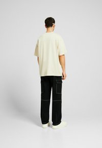 Muž v béžovém oversized tričku, černých cargo kalhotách s bílým prošíváním a bílých teniskách, stojící čelem ke světle šedé zdi.