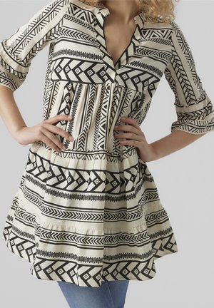 Vrouw draagt een zwart-wit jurk met geometrische patronen en driekwartmouwen, handen op de heupen tegen een neutrale achtergrond.