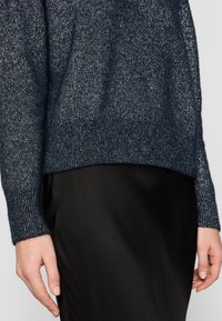 Maglione lavorato a maglia blu scuro con fili metallici, con polsini e orlo a coste, abbinato a una gonna nera elegante. Texture morbida e vestibilità rilassata.