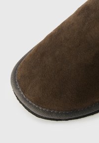 Καφέ σουέτ παντόφλες με στρογγυλή μύτη και αντίθετο μαύρο ύφασμα γύρω από τη σόλα, με υφή που προσφέρει πρόσφυση.