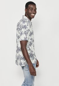Camicia a maniche corte in tessuto bianco con motivo a foglia di palma blu; abbinata a jeans azzurri chiari, modello in piedi di lato e sorridente.