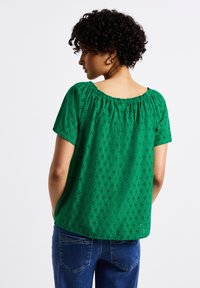 Top verde a maniche corte con scollo elasticizzato, caratterizzato da motivo ad occhielli. Indossato con jeans blu, mostrato da un'angolazione laterale.