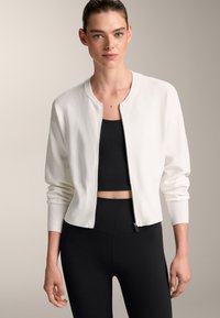 Bela bomber jakna z ribastimi manšetami in obodom, z zadrgo spredaj. Oblačena čez črn crop top in črne hlače z visokim pasom. Gladka tekstura.