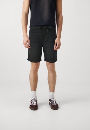 Mand iført sorte shorts med snøre, hvide sokker og mørkerøde sneakers står mod en ensfarvet lys baggrund.