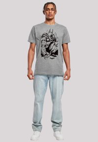 Grå bomulls t-skjorte med en svart grafikk av en superhelt. Modellen har på seg lyseblå denimjeans og hvite sneakers.