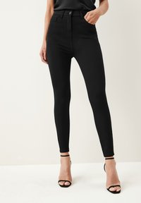 Černé skinny džíny s vysokým pasem vyrobené z elastického materiálu, s knoflíkovým zapínáním, poutky na pásek a kapsami, doplněné černými páskovými podpatky.