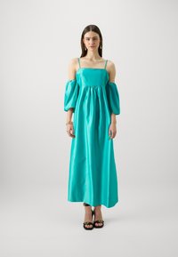 Akvakék selyem maxi ruha puffos ujjal, négyzet alakú dekoltázzsal és gyűrt derékkal. A modellen fekete szandál, minimális kiegészítőkkel.