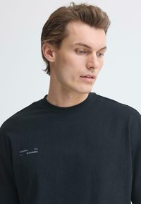 T-shirt noir à manches longues en coton, avec un col rond et un texte imprimé en gris : « La raison d'être vous ancre. » Texture lisse.