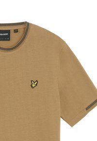 Brun bomullströja med korta ärmar, ribbad mörk krage, och en liten gul och svart broderad logotyp på bröstet.