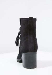 Bottine en suède noir avec un talon empilé, dotée d'une fermeture éclair sur le côté et de franges décoratives à l'arrière pour plus de détails.