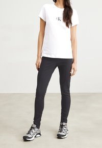 Weißes Baumwoll-T-Shirt mit schwarzem Logo, kombiniert mit enganliegenden schwarzen Hosen und grauen Sportschuhen mit schwarzen und silbernen Akzenten.