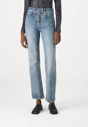 Jeans bleu clair à coupe droite avec fermeture zippée et bouton sur le devant, portés avec des mocassins noirs et un haut à manches longues à motifs.